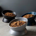 Gamelles inclinées Esprit Chat en céramique pour chat, disponibles en gris, noir et bleu, remplies de croquettes.