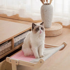 Chat Ragdoll assis sur un hamac Esprit Chat en bois avec toile rose et beige, installé près d'un meuble en bois.
