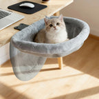 Hamac pour chat Esprit Chat en fausse fourrure grise, fixé au bord d'un bureau en bois avec un chat assis dedans.